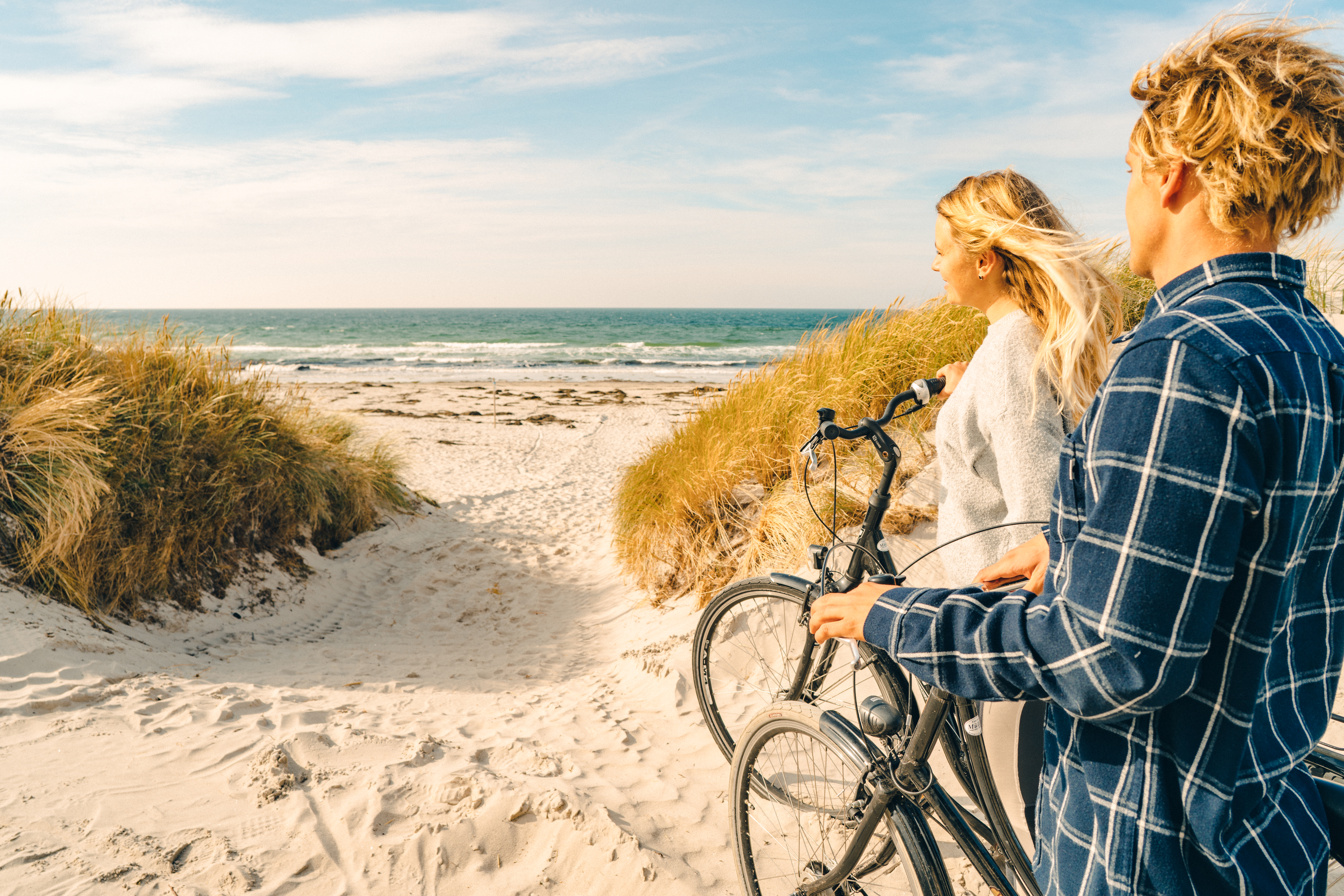 Auf dem Bild sieht man den Blick aufs Meer auf der Insel Hiddensee durch die Dünen. Rechts im Bild stehen ein Mann und eine Frau mit dem Rücken zur Kamera, die ihre Fahrräder Richtung Meer schieben.